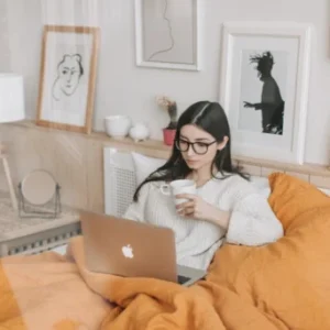A woman sits in bed with orange bedding, using a laptop and holding a cup.